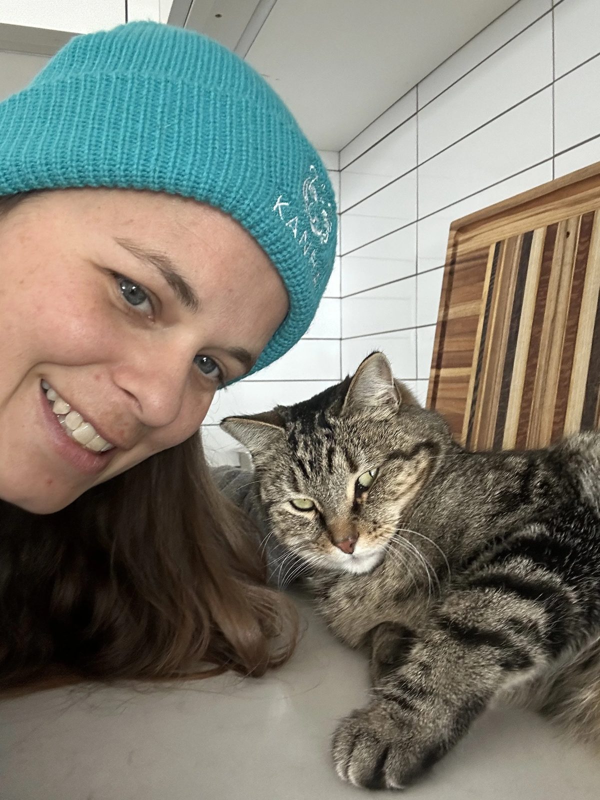 Lauren in a teal beanie smiling beside a senior tabby cat resting on a kitchen counter