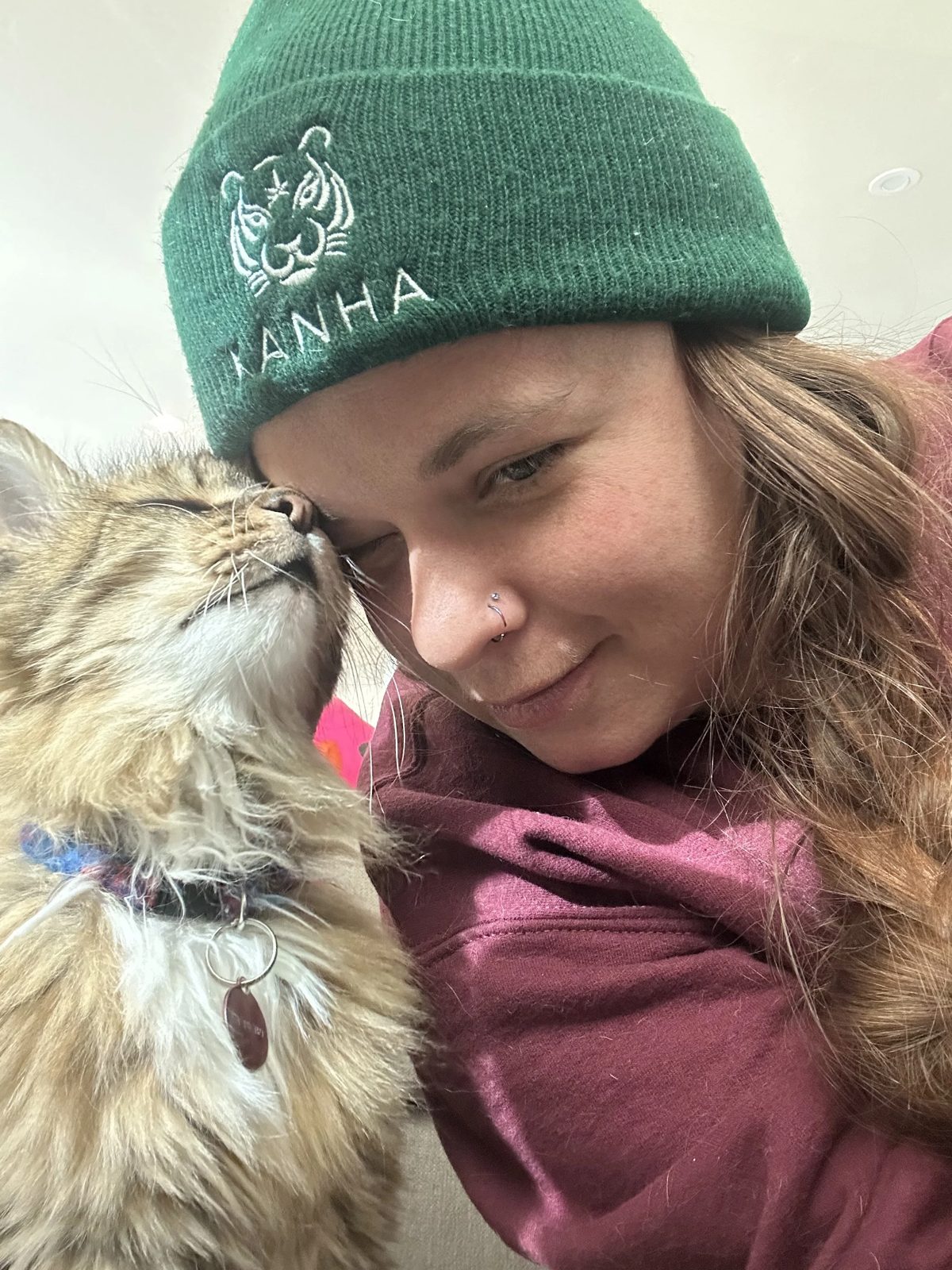 Lauren in a green beanie touching foreheads with a long-haired tabby cat who has its eyes closed
